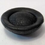 Image: A black button-shaped object made of natural glass. It is roughly circular, and features a convex face. The opposite face comprises a shallow depression with a central convex surface emerging from it