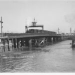 Image: A steam train crosses a bridge that extends across a narrow river. Several buildings are visible along the waterfront in the background