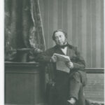 Image: a man sits cross legged on a chair, holding a newspaper