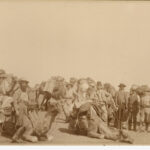 Image: ten camels, all with luggage and saddlebags tied to them with rope, and eight men preparing for an expedition