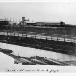 Image: A large paddock, behind which is a large complex of long, single-storey late Victorian-era stone buildings with steam trains parked in front of them. A two-storey building and several ship masts are visible in the distant background