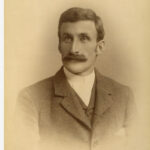 Image: A photographic head-and-shoulders portrait of a young Caucasian man wearing a late-Victorian era suit (without tie). He is sporting a large, luxurious handlebar moustache