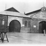 Image: A woman walks by two long brick buildings, one of which has the words ‘Edwin Daw Central Fish Market’ on its facade