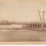 Image: A large iron and wood bridge under construction. Both approaches are nearing completion, while the central span is yet to be started. A barge with cranes is moored in the river near the bridge