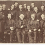 Image: A group of Caucasian men in mid-Victorian Catholic clergymen outfits pose for a photograph in a studio. The younger men in the group stand behind four older men who are seated