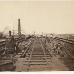 Image: A construction crew works on a bridge in a nineteenth-century port. A hard-hat diver and two well-dressed men are seated at centre right. Tall ships are visible in the adjacent river