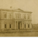 Image: a two storey stone building with a columned portico and low pitched roof