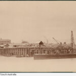 Image: An iron and wooden bridge under construction extends across a narrow river. A nineteenth century sailing vessel and a couple of buildings are visible in the background. A wooden-hulled barge with a derrick is moored next to the bridge