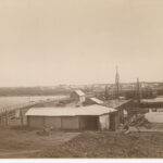 Image: A bridge under construction extends across a narrow river. A narrow wooden footbridge is located to the left of the bridge that is being built. Several nineteenth century buildings are visible in the distant background