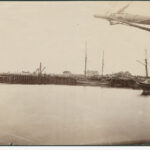 Image: The approaches to a large wooden and iron bridge are in the process of being built. A section of river containing a number of nineteenth century sailing ships are visible in the right foreground