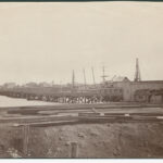 Image: A wooden footbridge extends across a river. A handful of nineteenth-century buildings and sailing ships are visible in the background