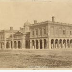 Image: A large, two-storey stone building in Victorian Italianate style. It features open archways on the ground floor, and its central section is topped by a cupola. A group of men stand in front of the left side of the building