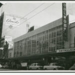 Image: a large 1950s era building with huge windows surrounded by much older buildings