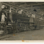 Image: The front of a train sitting in a large shed with five people sitting on the frond looking towards the camera