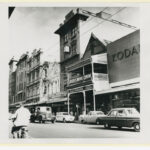 Image: 1960s cars (and a man on a bicycle) pass in front of a row of much older buildings. One of these, the Globe Theatre, is particularly ornate.