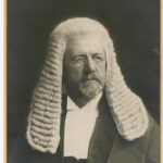 Image: A photographic head-and-shoulders portrait of a bearded middle-aged Caucasian man wearing a judicial wig and vestments. He is also wearing wire-rimmed spectacles and looking to his left