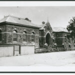 Image: a single-storey slate-roofed bluestone Gothic style building with a gabled front entrance and arched windows