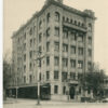 Image: a six storey reinforced concrete building with a stone façade. It features bay windows with balconies on the first and sixth floors.  An awning runs down one side of the ground floor with large shop windows visible underneath.