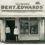 Image: Two men stand in the doorway of a small shop building. A sign on the roof of the shop reads ‘Tea Rooms, Bert.Edwards’