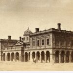 Image: A large, two-storey stone building in Victorian Italianate style. It features open archways on the ground floor, and its central section is topped by a cupola