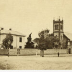 Image: A dark stone church with a lighter stone square tower at one end sits beside a cream coloured square two-storey residential building behind a stone wall.