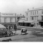 Image:  A three horse team and laden wagon is standing on a dirt street near an intersection. In the centre of the view, a man on a scaffold is engaged in building work. The building on the near side of the intersection has a sign reading "The Bee Hive"