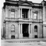 Image: two men in suits pose on the front steps of a two-storey stone building with recessed main entrance flanked by stone columns which are repeated on the floor above framing a balcony and topped with a simple triangular pediment.