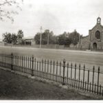 Image: A large stone church and stone house stand adjacent to one another next to a dirt street. Part of an iron fence is visible in the immediate foreground