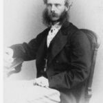 Image: Black and white portrait of a partially balding man with long mutton chops and a moustache sitting in a chair. He is wearing a shirt and suit jacket