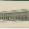 Image: a long two storey brick building with clerestory windows rising from a low pitched roof and with arched windows and doors on the main body of the building.