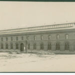 Image: a long two storey brick building with clerestory windows rising from a low pitched roof and with arched windows and doors on the main body of the building.