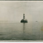 Image: A metal lighthouse with a large wooden support platform stands surrounded by water. Large steam- and sail-powered ships are visible in the distant background