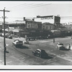 Image: Various mid-twentieth century auto-mobiles pass through an intersection in an urban area. A large plinth stands in the centre of the intersection and is marked with the words ‘Keep Left’ and left-facing arrows