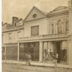 Image: A man and a boy in 1870s clothing stand outside a row of two storey terraced shops. The man is leaning on a post and the boy stands behind him.