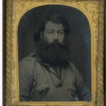 Image: a head and shoulders portrait of a man with long dark hair and a very thick long black beard. He is wearing a casual 1850s era shirt.