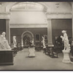 Image: The interior of an art gallery with high ceilings. Six white statues and numerous portrait paintings are in view