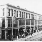 Image: a large three storey building with full height shop windows on the ground and first floors displaying a range of merchandise. A large number of people are walking past on the sidewalk.