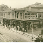 Image: a city department store, mostly two storeys but with one three storey section. Men and women in early 20th century clothes are peering in the plate glass windows at the displays.