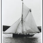 Image: A small, single-masted sailing vessel with a cutter rig coasts off a nearby shoreline. A crew of approximately a dozen men are visible standing along the vessel’s length