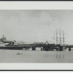 Image: A steam train crosses a bridge that extends across a narrow river. A large nineteenth century sailing vessel is visible behind the bridge, while a small rowboat operated by two men is present in the foreground