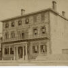 Image: A three storey brick and stone building with shuttered windows and a balcony running the length of the second floor above a columned portico.