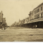 Image: a wide dirt city street, lined with shops, with a single horse drawn vehicle parked on the left of the street. Men are posing for the photograph on both sides of the road outside of shops.