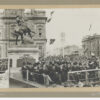 Image: a group of men in dark suits and top hats sit on a stage in front of a statue of a soldier on a horse. A large crowd surrounds them.