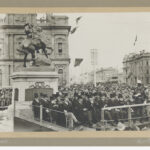 Image: a group of men in dark suits and top hats sit on a stage in front of a statue of a soldier on a horse. A large crowd surrounds them.