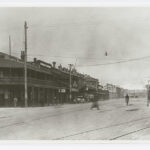 Image: Several Victorian-era buildings line a dirt street criss-crossed by railway tracks. A handful of people and cars of 1920s vintage are visible on the street and adjacent sidewalk