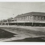 Image: An intersection of two streets in an urban area. A plinth with the words ‘Keep Left’ stands in the centre of the intersection. Early twentieth century cars and a horse and cart are parked in the background