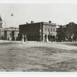 Image: An intersection of two streets in an urban area. A plinth with the words ‘Keep Left’ stands in the centre of the intersection. Two early twentieth century cars are parked in the background