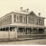 Image: A large, two-storey stone building bounded by a white fence and gate