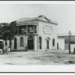 Image: Men, women and children gather in front and on the balcony of a small hotel, painted white with a corner door and the name "Hampshire Hotel" surrounded by a decorative border on the side.