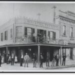 Image: group stand in front of a tavern building with four women standing on a balcony in the tavern